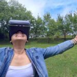 Woman wearing VR glasses with arms open wide and smile on green grass surrounded by trees.