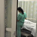 Image shows doctor attending patient in hospital bed with back to camera through partially open doorway