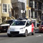 Image of small white and red vehicle with camera and navigational equipment on roof driving through street in San Francisco