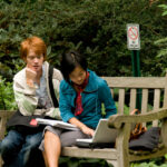 Young adults reading on a laptop on a bench outdoors.