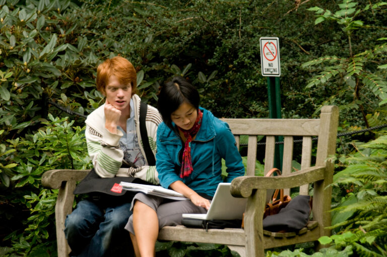 Young adults reading on a laptop on a bench outdoors.